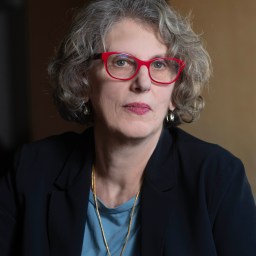 A woman with short curly gray hair and red glasses wearing a blue top, black blazer, gold earrings, and a necklace with a star-shaped pendant, poses in front of a neutral background.