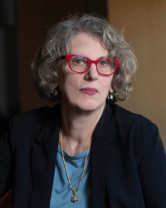 A woman with short curly gray hair and red glasses wearing a blue top, black blazer, gold earrings, and a necklace with a star-shaped pendant, poses in front of a neutral background.