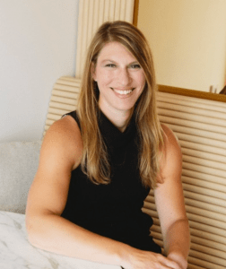 A woman with long blonde hair wearing a sleeveless black top sits at a table, smiling at the camera.