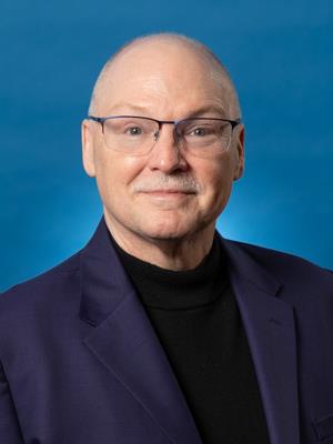 A middle-aged man with glasses, short light hair, and a mustache, wearing a dark blazer and black turtleneck, poses in front of a blue background.