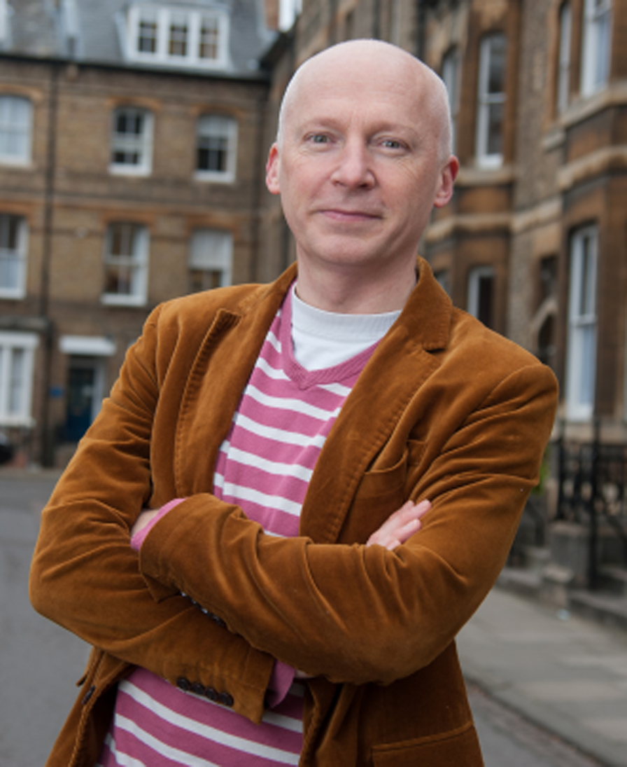 A person with a shaved head wearing a brown corduroy jacket and a pink striped shirt stands outdoors with arms crossed in front of brick buildings.