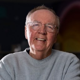 An older man with glasses, short hair, and a light gray sweater smiles while sitting indoors with a dark background.
