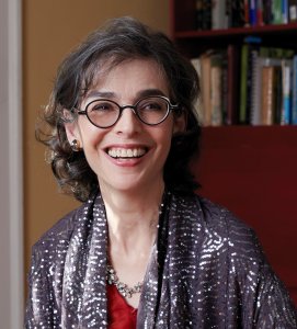 A woman with glasses and curly hair, smiling, wearing a sequined shawl and a red top, seated in a room with a bookshelf in the background.