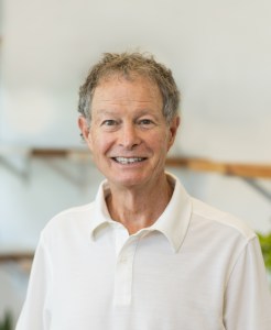 A smiling man with short, curly gray hair wearing a white collared shirt stands in an indoor setting.