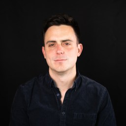 A man with short dark hair wearing a dark button-up shirt poses against a plain black background.
