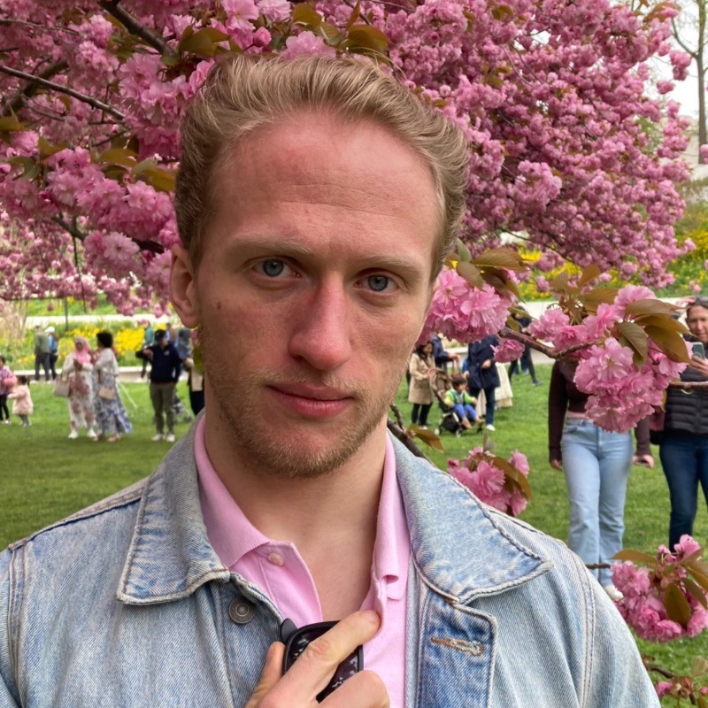 Person with light hair and a denim jacket stands in front of pink cherry blossoms in a park. People and green grass are in the background.
