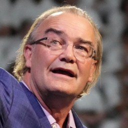 An older man with glasses and gray hair, wearing a blue suit jacket, is speaking. He looks slightly upwards, with a background of blurry, out-of-focus shapes.