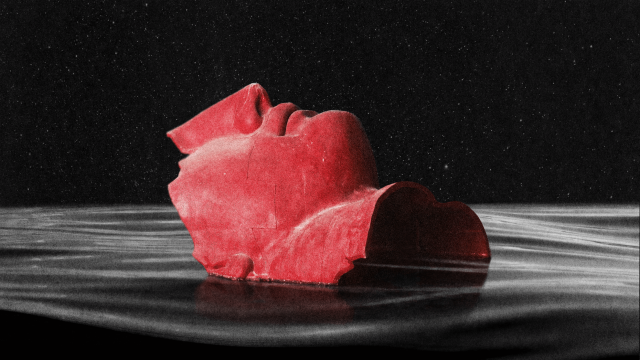 A red, fragmented sculpture of a human face is partially submerged in dark, rippling water against a black background.