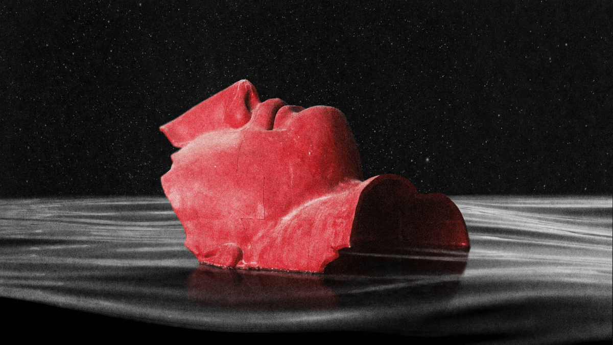 A red, fragmented sculpture of a human face is partially submerged in dark, rippling water against a black background.