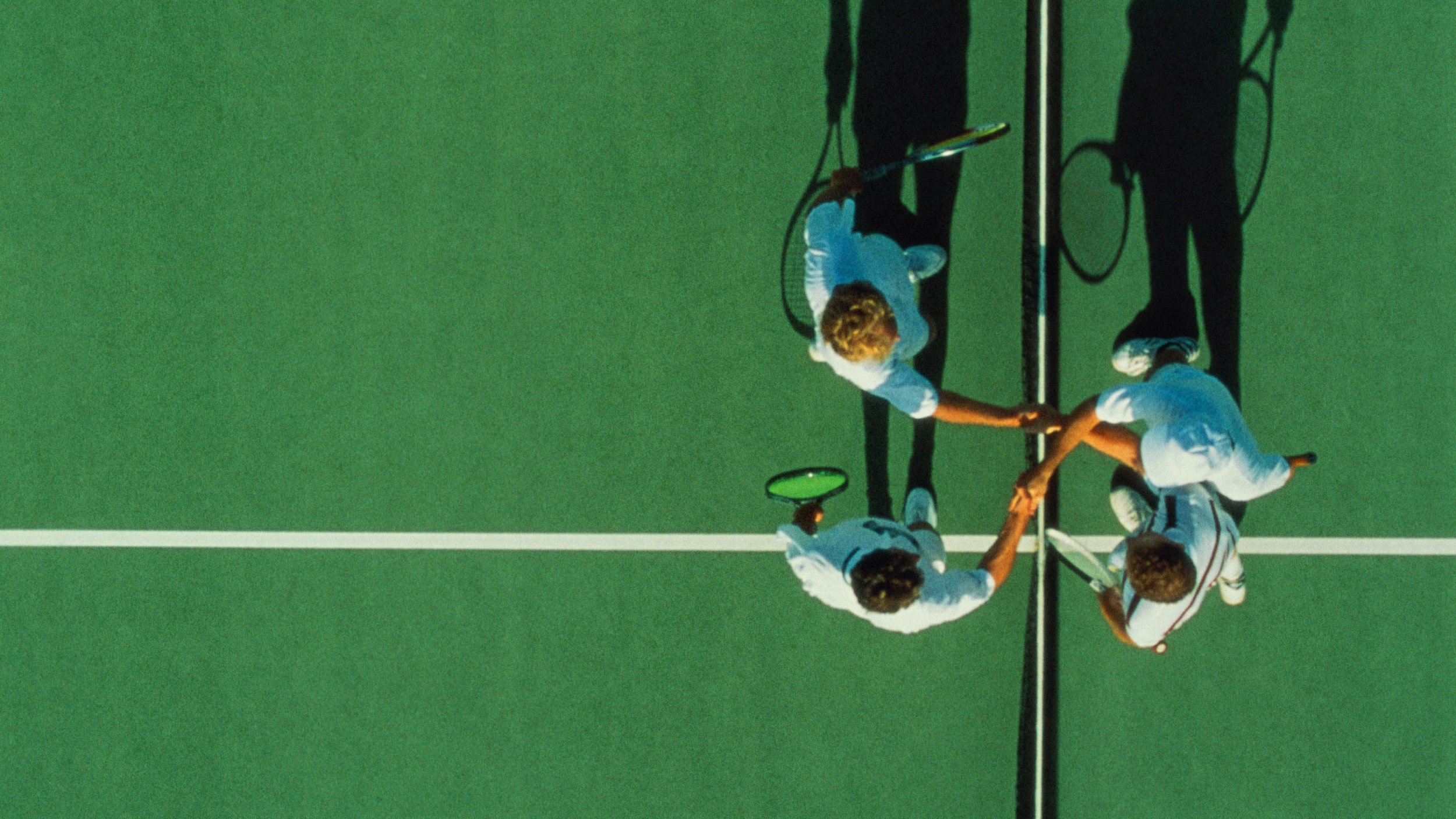 An aerial view captures two pairs of tennis players, akin to The Four Enemies, shaking hands at the net on a lush green court.