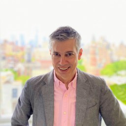 a man in a suit and pink shirt standing in front of a window.