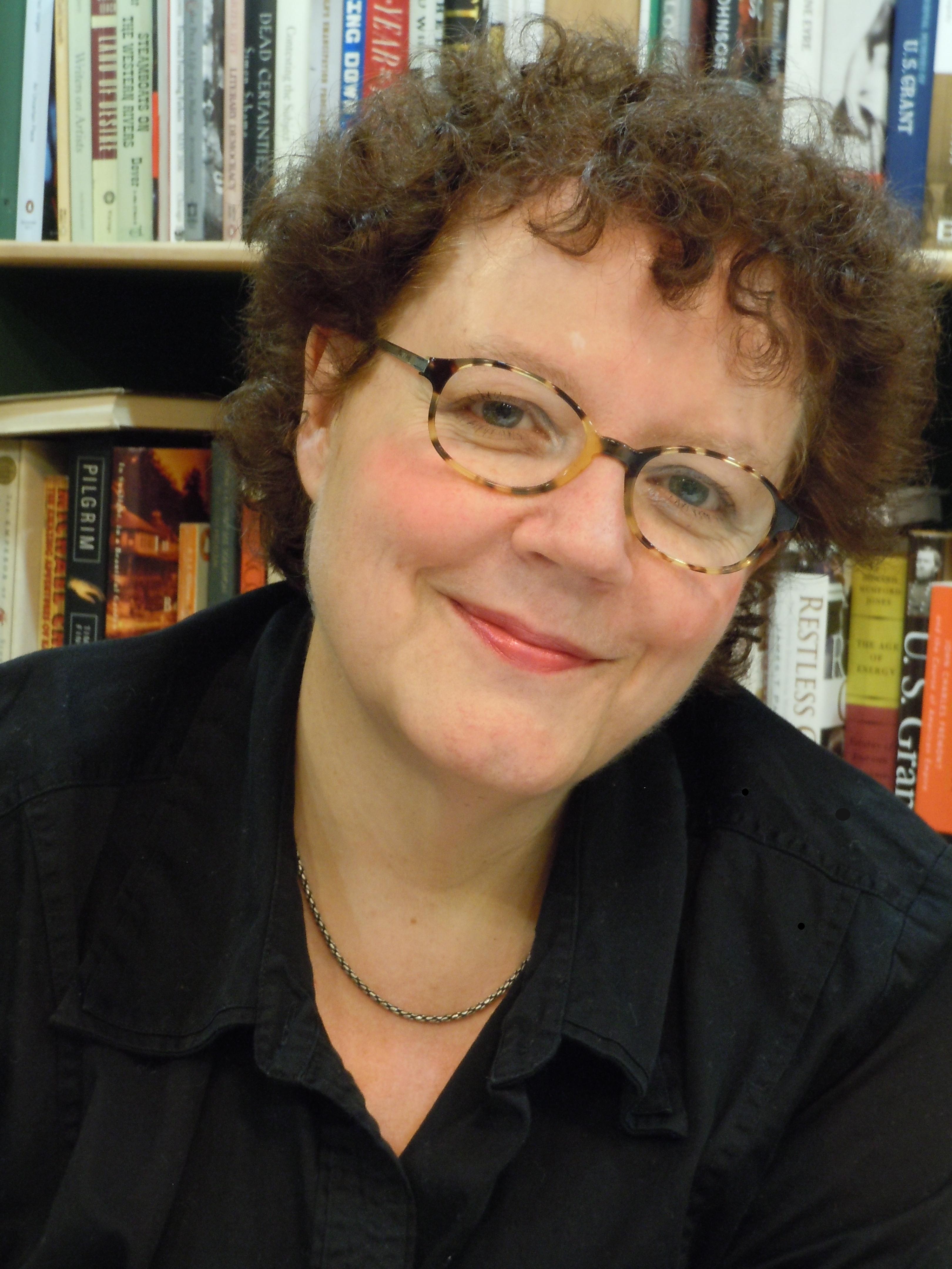 a woman with glasses smiling in front of a bookshelf.