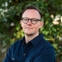 A man in glasses standing in front of bushes.