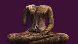 Ancient headless stone Buddha statue with one arm missing, seated cross-legged against a solid dark purple background.