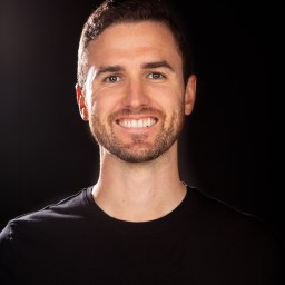 A man in a black shirt smiling in front of a black background.