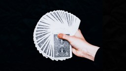 A hand holds and fans out a deck of playing cards with patterned backs against a black background.