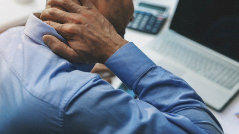A man in a blue shirt is holding his neck in front of a laptop, possibly experiencing inflammation.