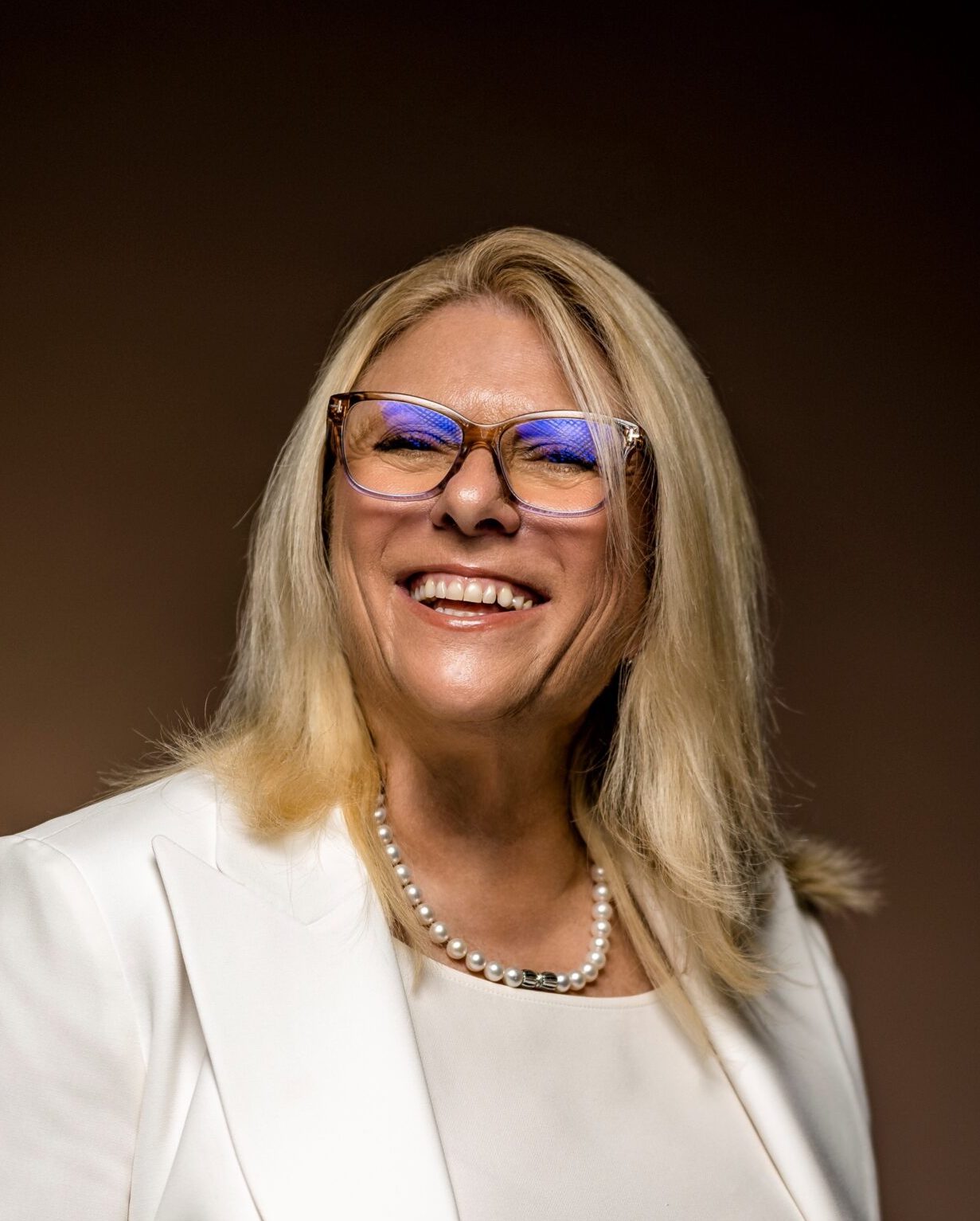 A smiling woman in glasses wearing a white blazer and pearls.