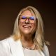 A smiling woman in glasses wearing a white blazer and pearls.