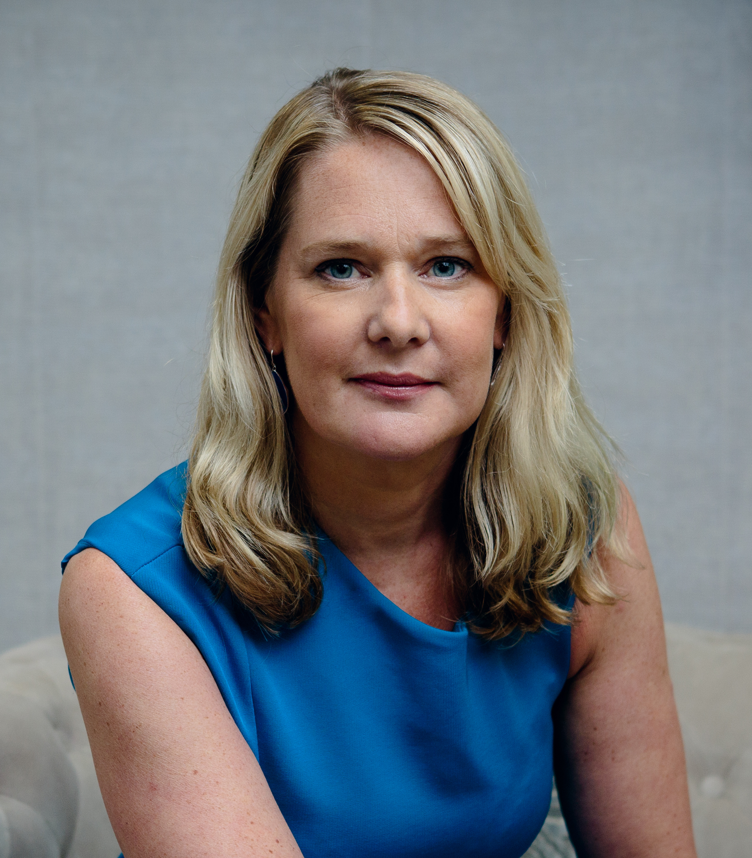 A woman with blond hair wearing a blue top poses for a portrait in front of a neutral background.