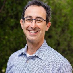 A smiling man with glasses wearing a blue patterned shirt, standing outdoors with green foliage in the background.