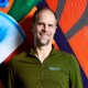 A smiling man with a beard in a green zip-up top stands before a colorful graffiti wall.