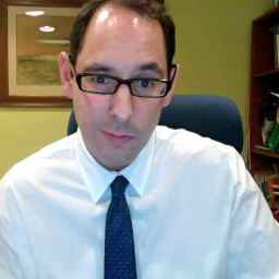 A man with glasses wearing a white shirt and tie, seated in an office with books and a framed painting in the background.