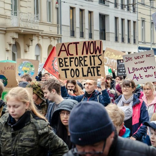 Crowd of diverse people at a climate protest holding signs with messages like "act now before it's too late".