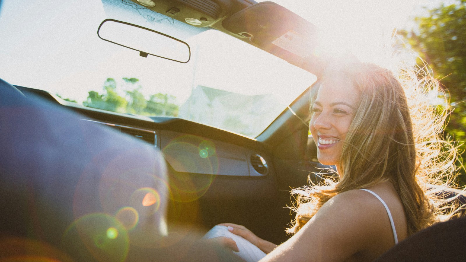 A smiling woman riding in a sunlit car, viewed from the passenger side, with windblown hair and sun flare visible.