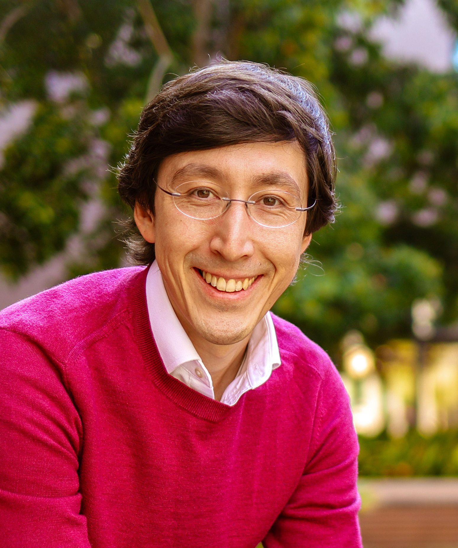 A person with glasses and dark hair, wearing a pink sweater over a white shirt, smiles while sitting outdoors with green foliage in the background.