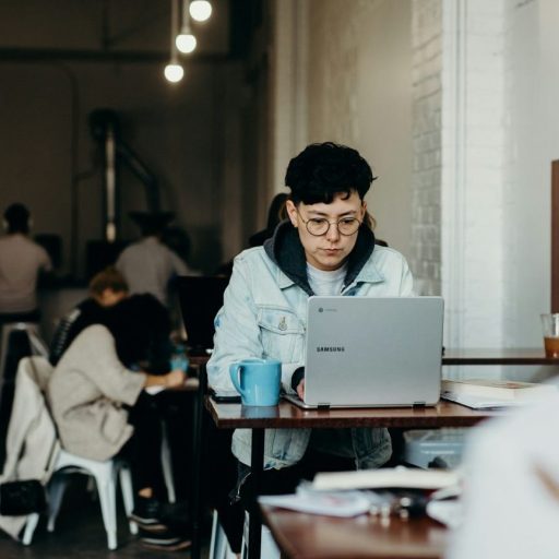 A person wearing glasses and a denim jacket works on a laptop at a table in a cafe. Several other individuals, including both humans and AI enthusiasts, are seated and engaged in different activities in the background.
