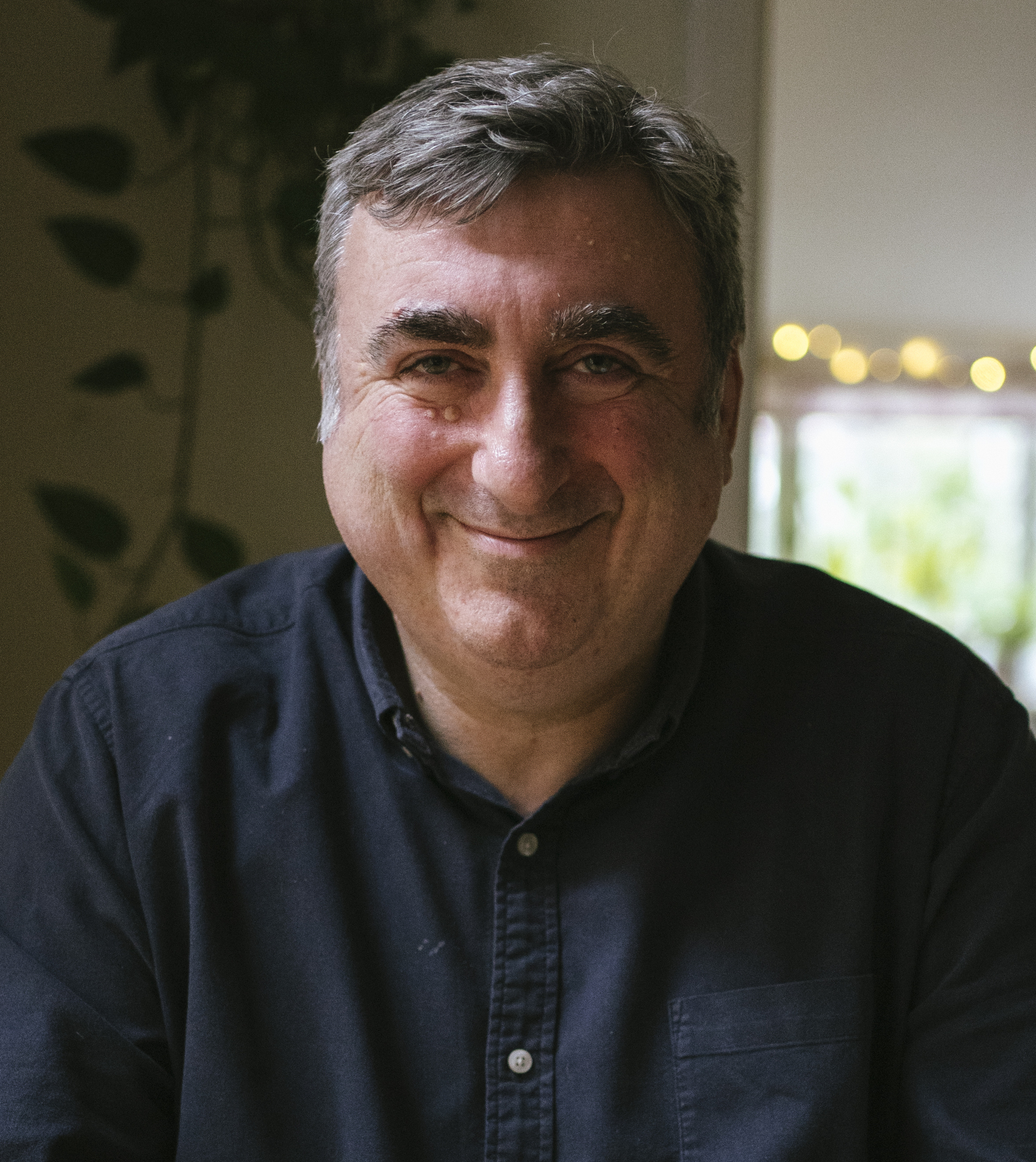 A middle-aged man with gray hair and a slight smile, wearing a dark button-up shirt, is sitting indoors with a plant and some string lights in the background.