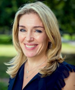 A woman with shoulder-length blonde hair, wearing a dark top and necklace, smiles in an outdoor setting.