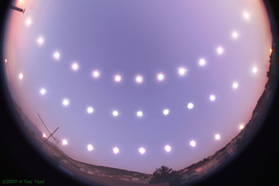 A time-lapse photo showing the Sun's path over several days, arranged to form a smiley face pattern against a twilight background. Taken around the period of the earliest solstice, &copy;2007-8 Tun&ccedil; Tezel is credited in the bottom left corner.