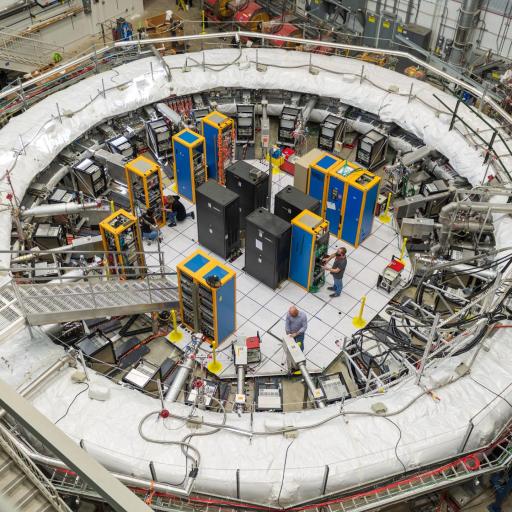 A large circular particle accelerator with several cables and machines is where engineers work inside and around the structure. The facility, dedicated to solving the muon g-2 anomaly, has platforms and specialized equipment surrounding the central structure.