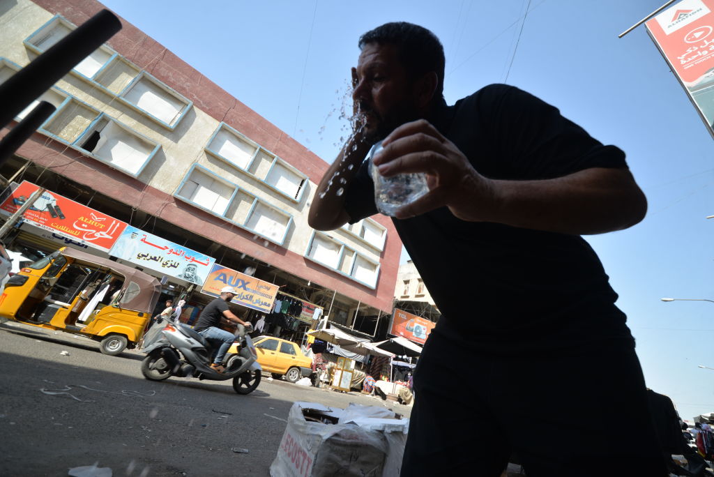 A man splashes water on his face from a plastic bottle on a busy street, with buildings, advertisements, and moving vehicles in the background.