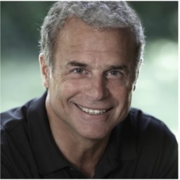 A man with short gray hair and a broad smile is wearing a dark polo shirt, looking directly at the camera against an out-of-focus green background.