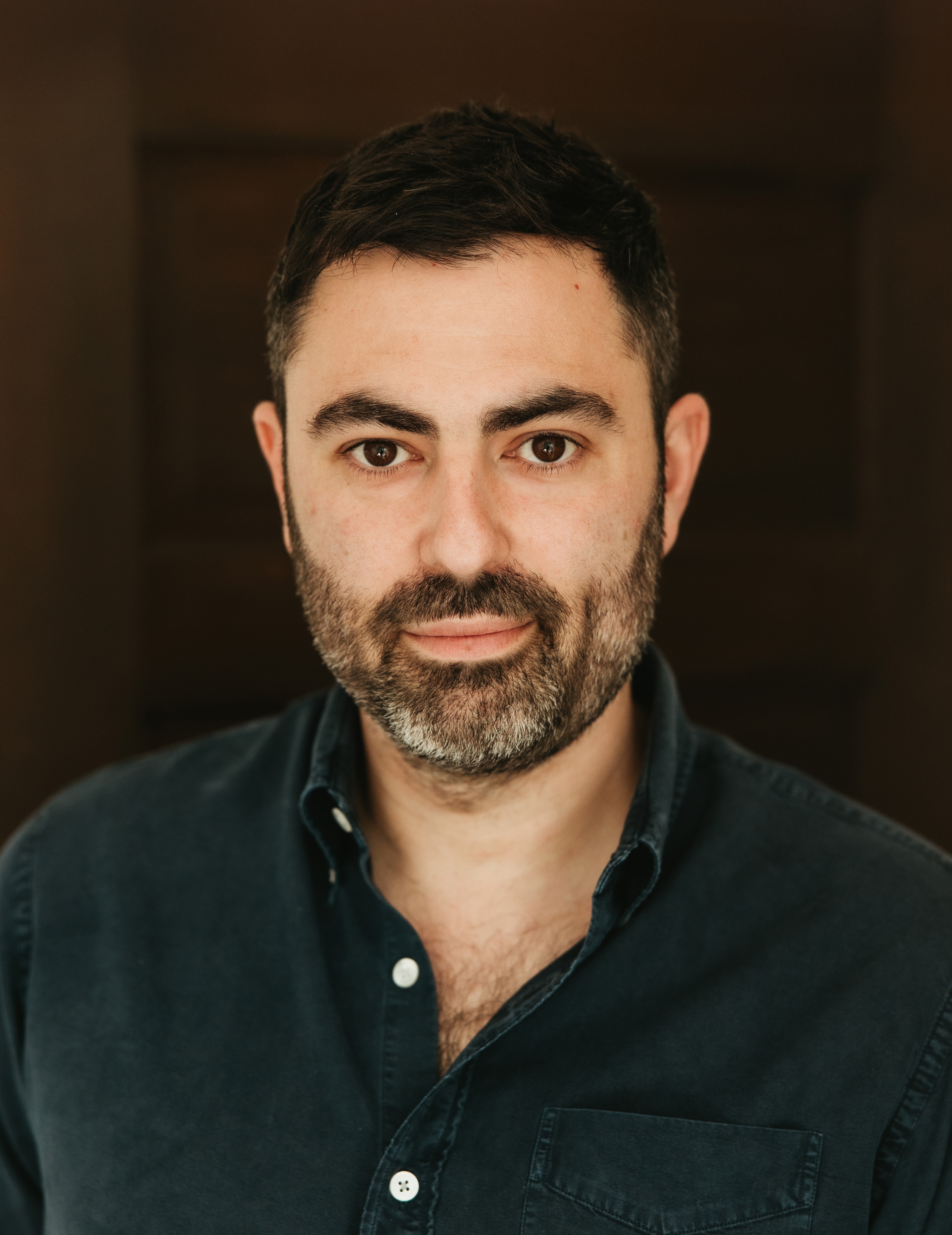 A man with short dark hair and a beard wearing a dark button-up shirt stands against a dark, blurred background, looking directly at the camera.