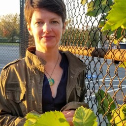 A person with short hair stands outside near a chain-link fence, holding green leaves with the sun casting a soft light.