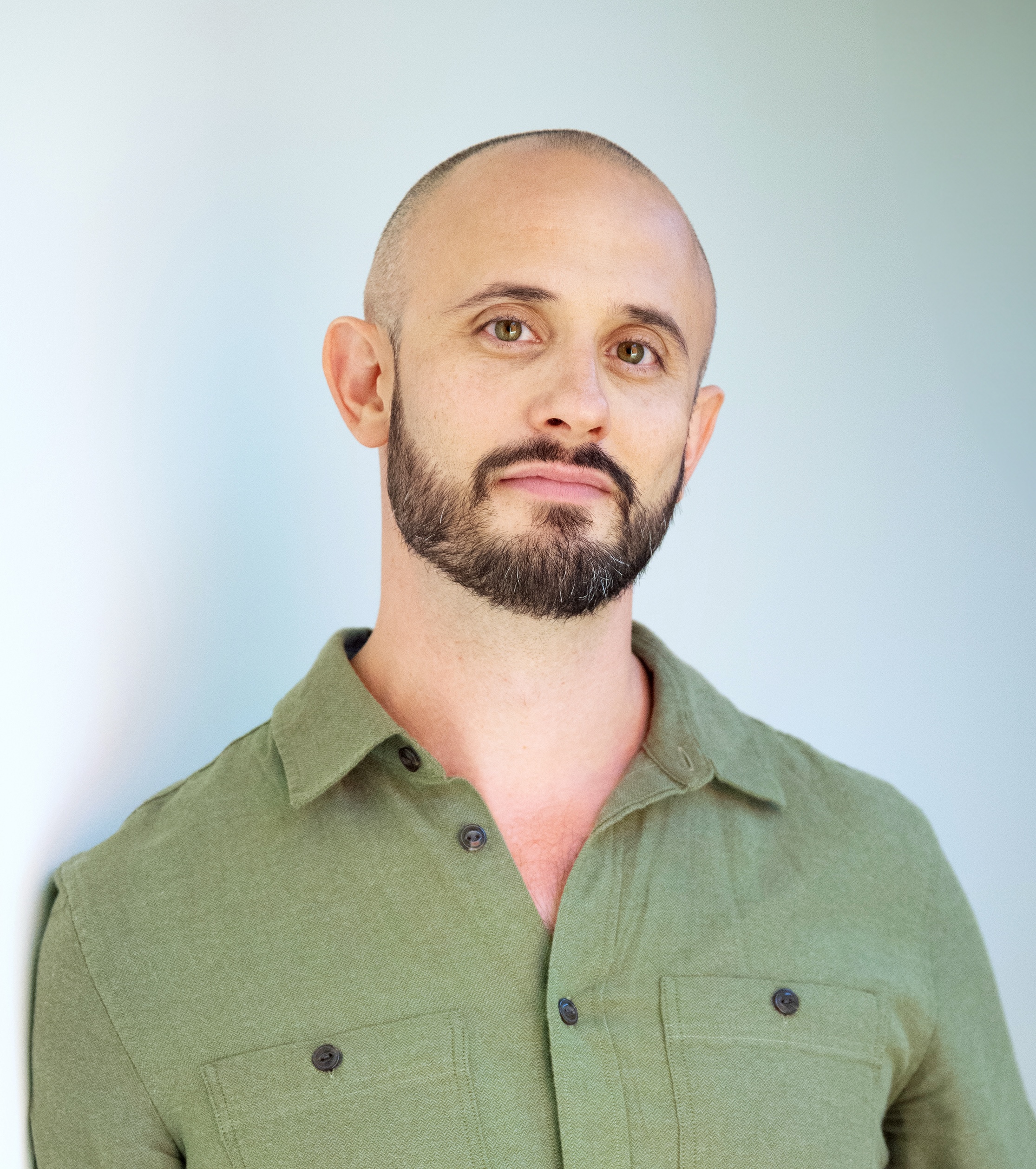 A man with a shaved head and a beard is wearing a green button-up shirt and standing against a plain background.