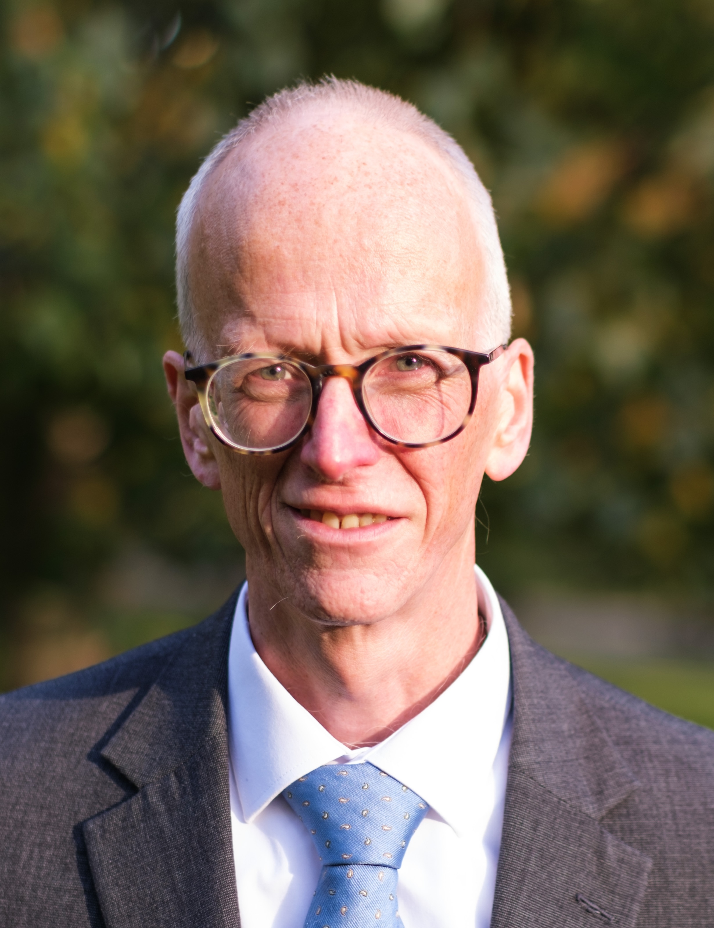 A bald man with glasses, wearing a gray suit, white shirt, and blue tie, stands outside with greenery in the background.