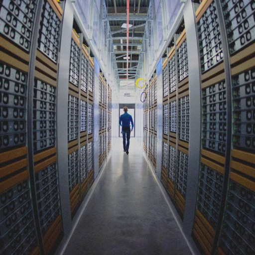 A wide shot of a man walking down an aisle between tall stacks of server racks in a data center filled with computer hardware.