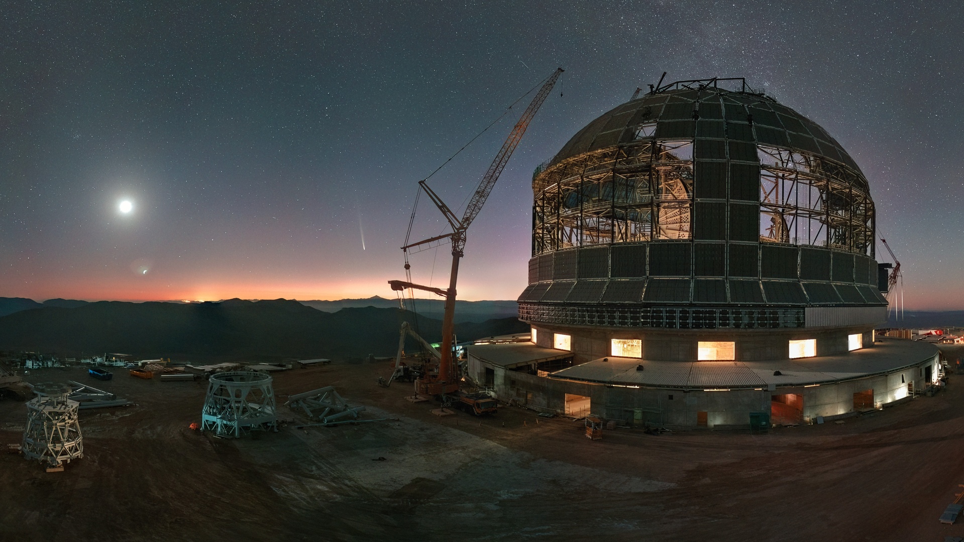 A large telescope observatory under construction at dusk with a visible moon and stars in the sky. Cranes and construction equipment are present around the structure.