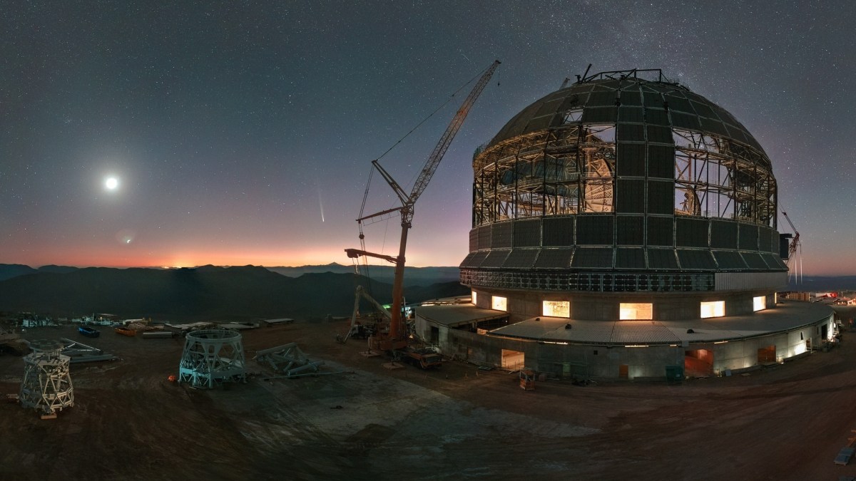 A large telescope observatory under construction at dusk with a visible moon and stars in the sky. Cranes and construction equipment are present around the structure.