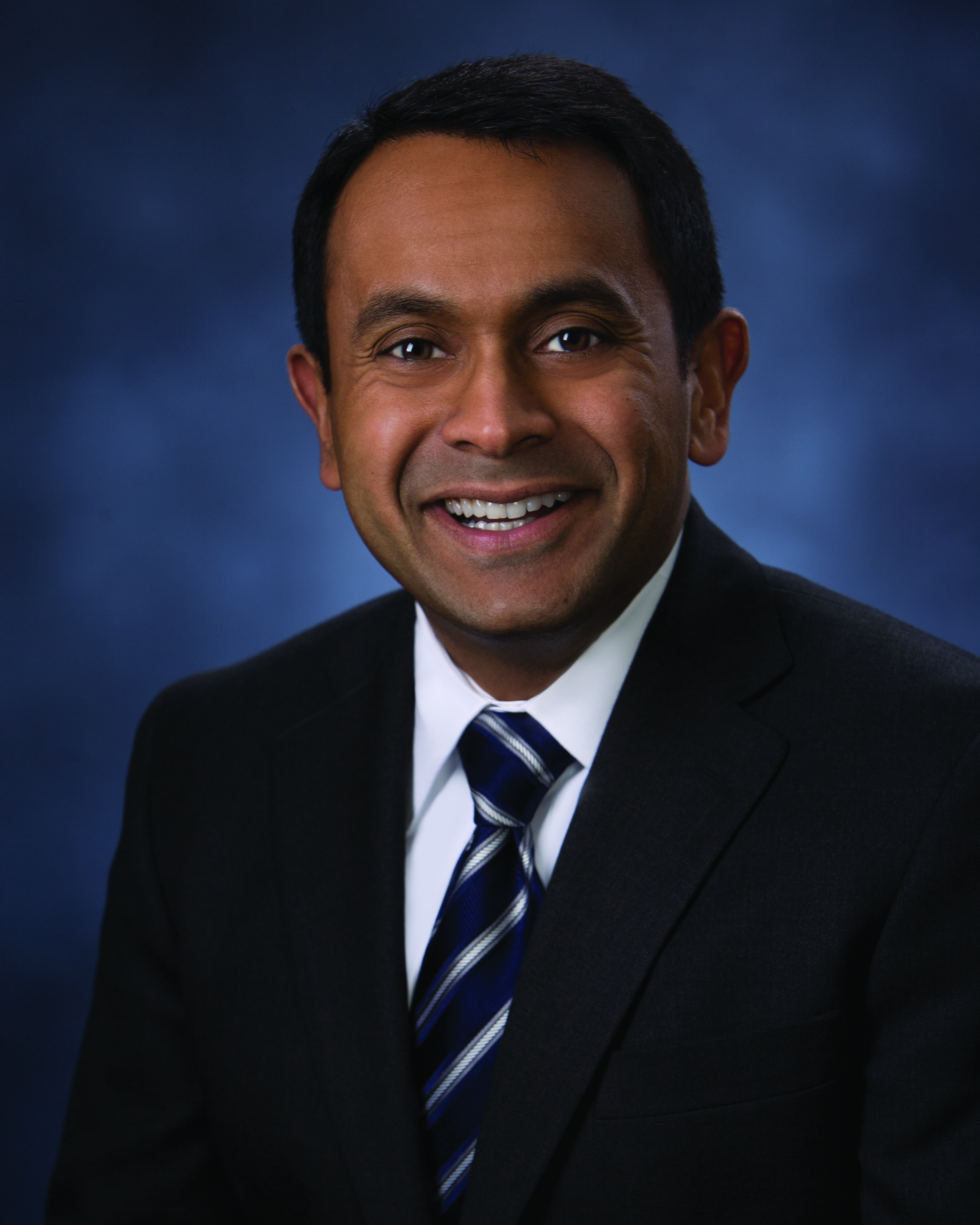 A man wearing a dark suit, white shirt, and striped tie is smiling against a blurred blue background.