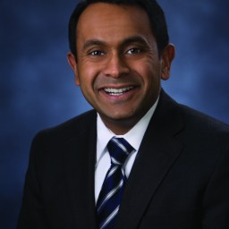 A man wearing a dark suit, white shirt, and striped tie is smiling against a blurred blue background.