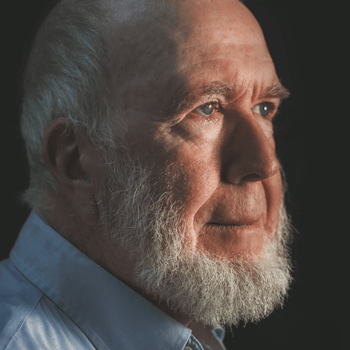 A side profile of an older man with a white beard, wearing a light blue shirt, against a dark background, illuminated by soft lighting from the left, exudes an aura of radical optimism.