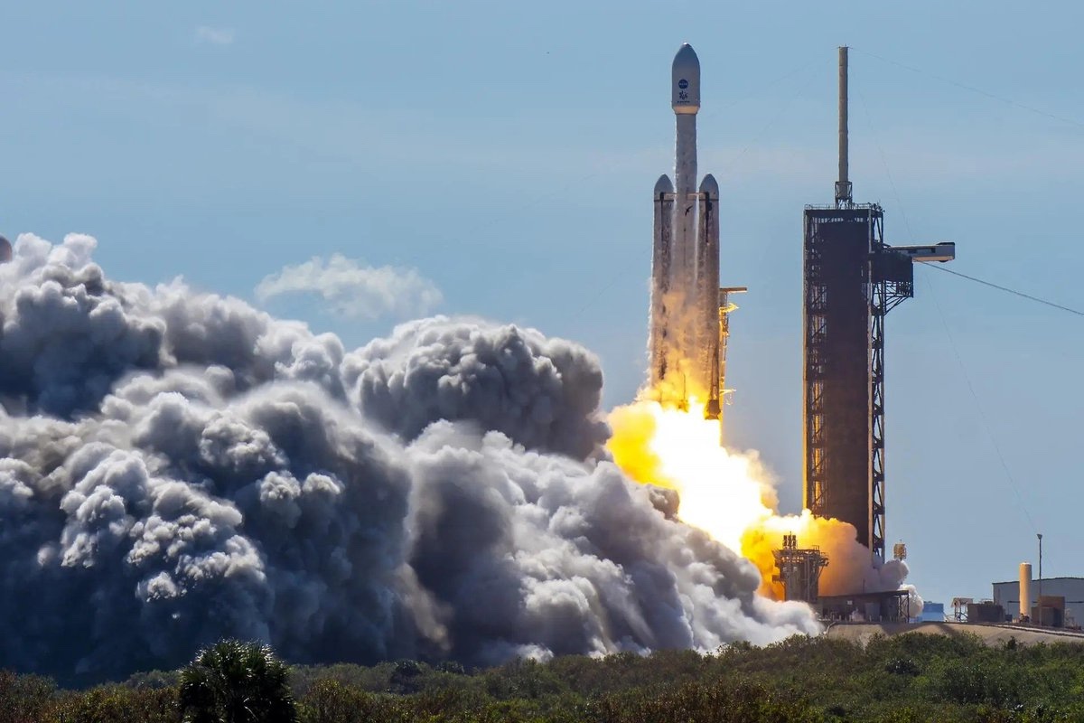 A rocket lifts off, generating dense clouds of smoke and flames, against a clear blue sky.