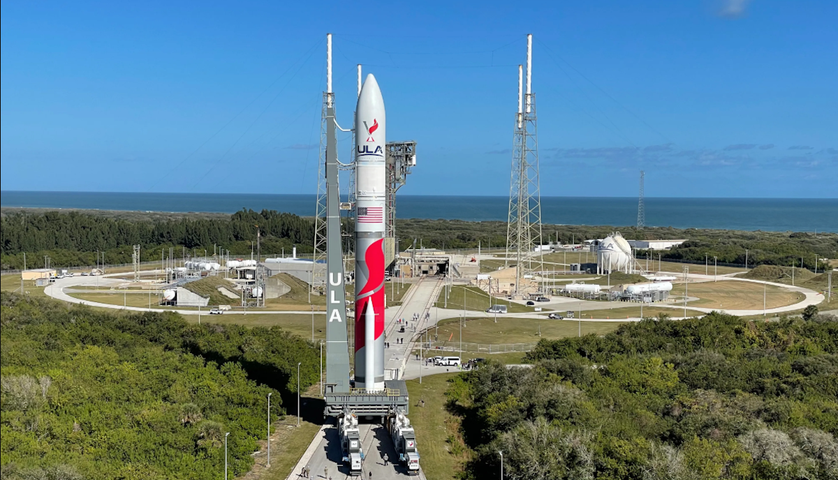 A rocket stands on a launch pad, surrounded by greenery and blue sky, with the ocean in the background.