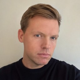 Portrait of a person with short light brown hair, wearing a black shirt, looking towards the camera with a neutral expression.